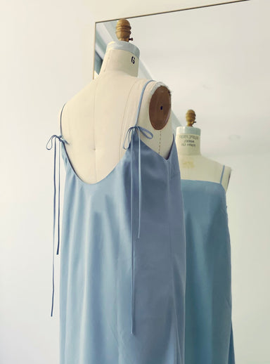 The Florence Lyocell Dress — Classic by Noémiah, a light blue slip dress with thin shoulder ties made from sustainable lyocell, is shown on a dress form in front of a mirror, displaying both front and back views in a bright, minimal room.