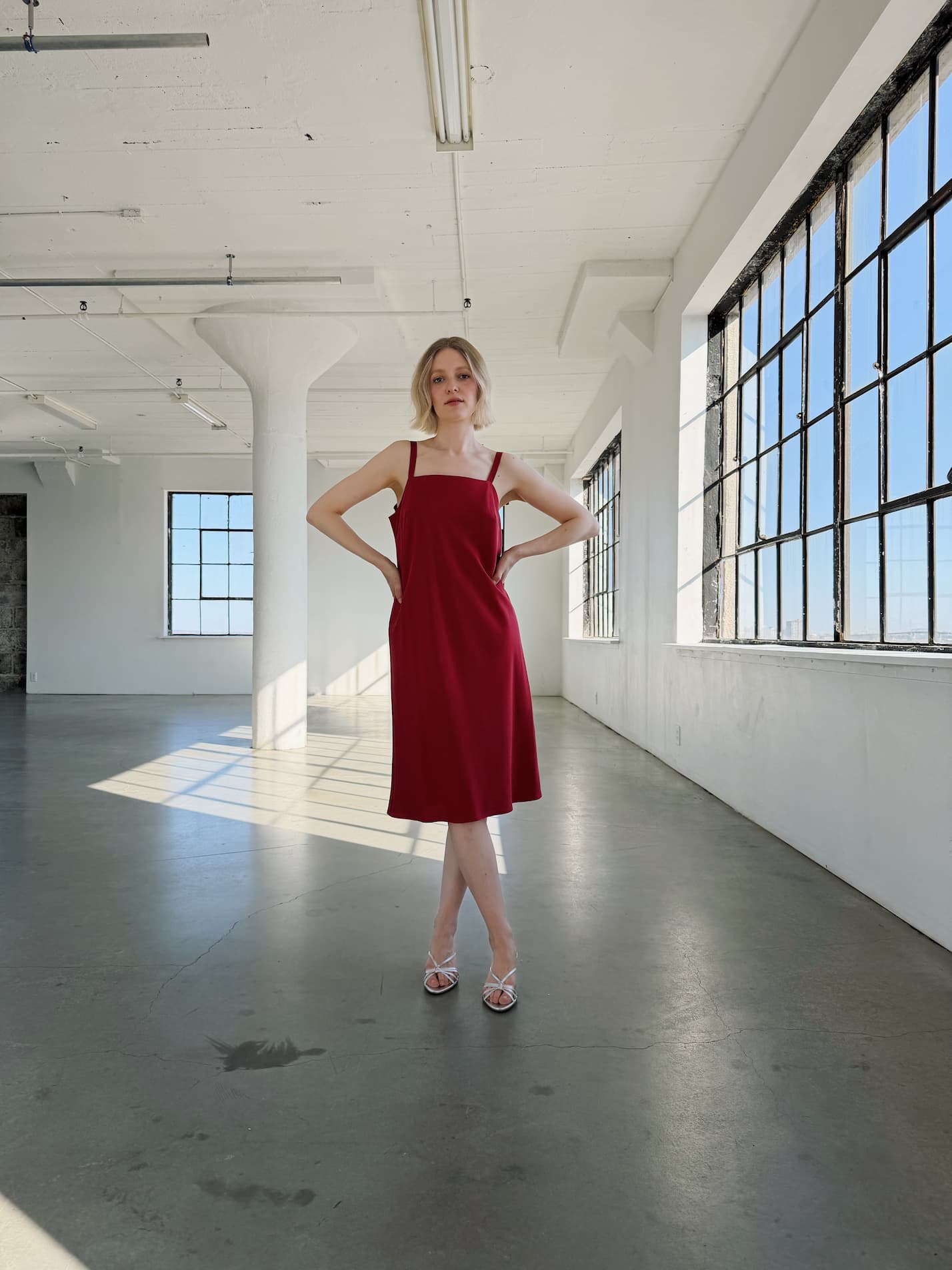 A woman in the Noémiah Florence Lyocell Dress — Classic, a sleeveless red sustainable slip dress, stands with hands on hips in a spacious sunlit room with large industrial windows and white walls.