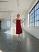 A woman in the Noémiah Florence Lyocell Dress — Classic, a sleeveless red sustainable slip dress, stands with hands on hips in a spacious sunlit room with large industrial windows and white walls.
