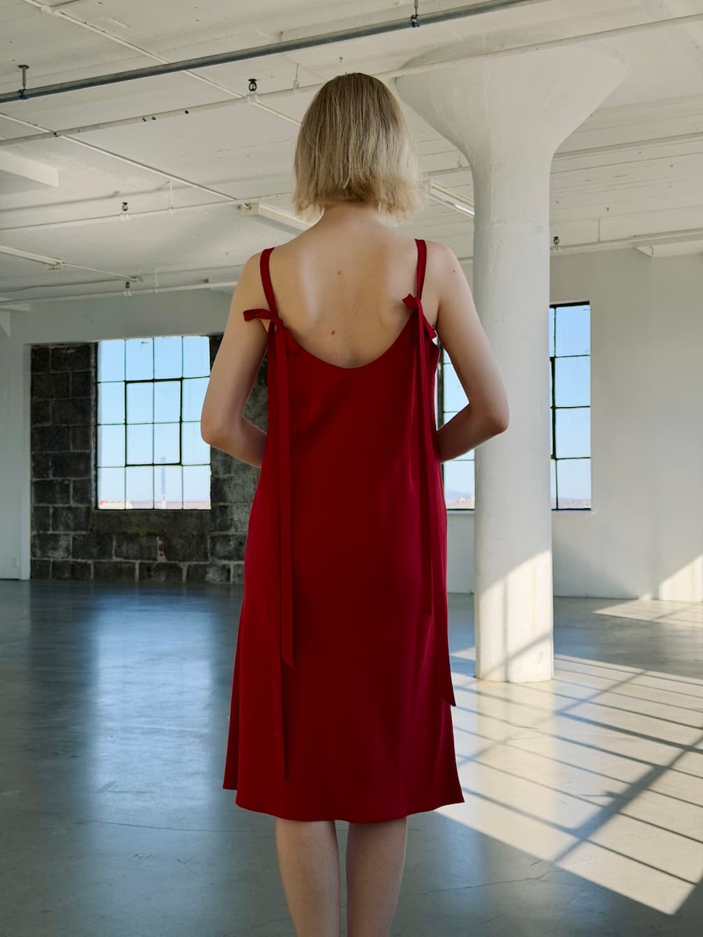 A person with short blonde hair stands in a sunlit, minimalist room wearing the Noémiah Florence Lyocell Dress — Classic, a sleeveless red slip dress with ribbon ties on the shoulders. Large windows light up the industrial space.