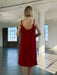 A person with short blonde hair stands in a sunlit, minimalist room wearing the Noémiah Florence Lyocell Dress — Classic, a sleeveless red slip dress with ribbon ties on the shoulders. Large windows light up the industrial space.