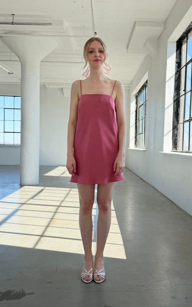 A woman stands in a sunlit, spacious room wearing the Noémiah Florence Lyocell Dress — Classic, a sleeveless pink slip dress made from sustainable Tencel, paired with white shoes. Soft shadows are cast on the floor by large windows.