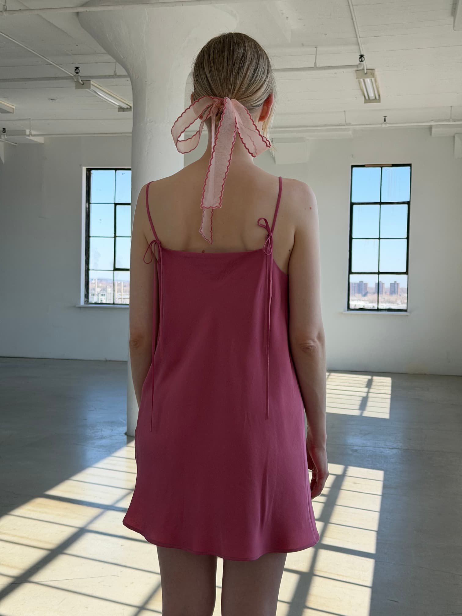 A woman stands indoors facing away, wearing the Noémiah Florence Lyocell Dress — Classic and a sheer organza hair ribbon, as sunlight streams through large industrial-style windows onto the floor.