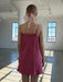 A woman stands indoors facing away, wearing the Noémiah Florence Lyocell Dress — Classic and a sheer organza hair ribbon, as sunlight streams through large industrial-style windows onto the floor.
