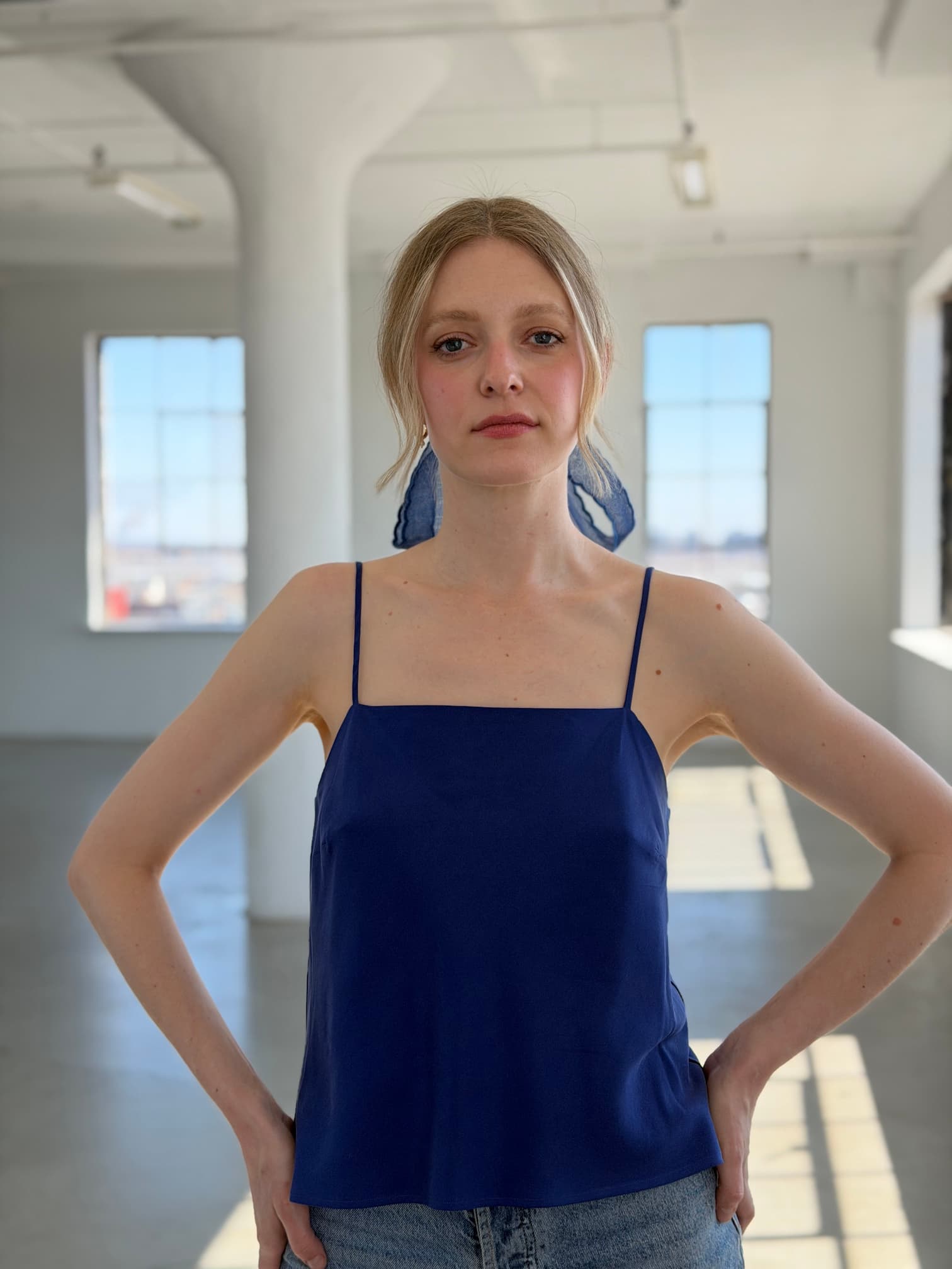 A woman with blonde hair tied back stands indoors, hands on hips, wearing Noémiah's blue sustainable lyocell Florence Top and jeans, in front of large windows filled with natural light.