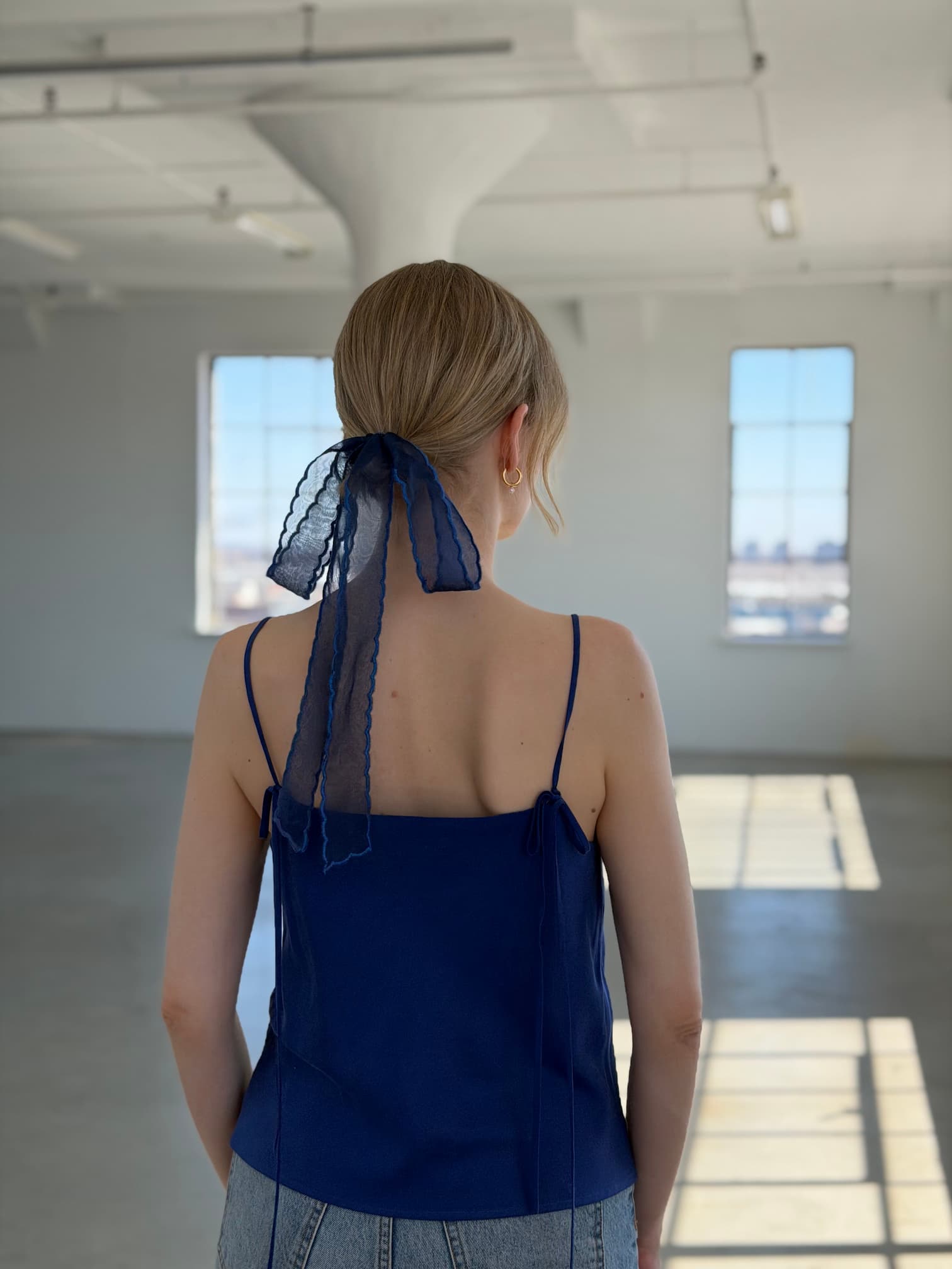 A woman stands in a sunlit room with large windows, her back to the camera. She wears Noémiah’s Florence Top in navy blue and has a sheer navy ribbon tied around her low ponytail of blonde hair.