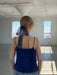 A woman stands in a sunlit room with large windows, her back to the camera. She wears Noémiah’s Florence Top in navy blue and has a sheer navy ribbon tied around her low ponytail of blonde hair.