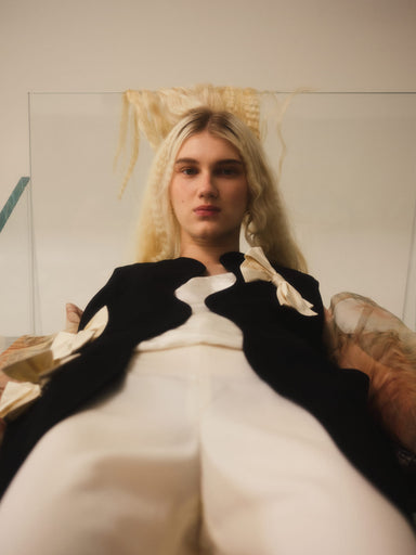 A person with long blonde hair is reclining and looking directly at the camera, adorned in the Françoise Vest by Noémiah. This tailored fit black and white outfit features large bows and a wool-crepe texture, set against a neutral indoor background.