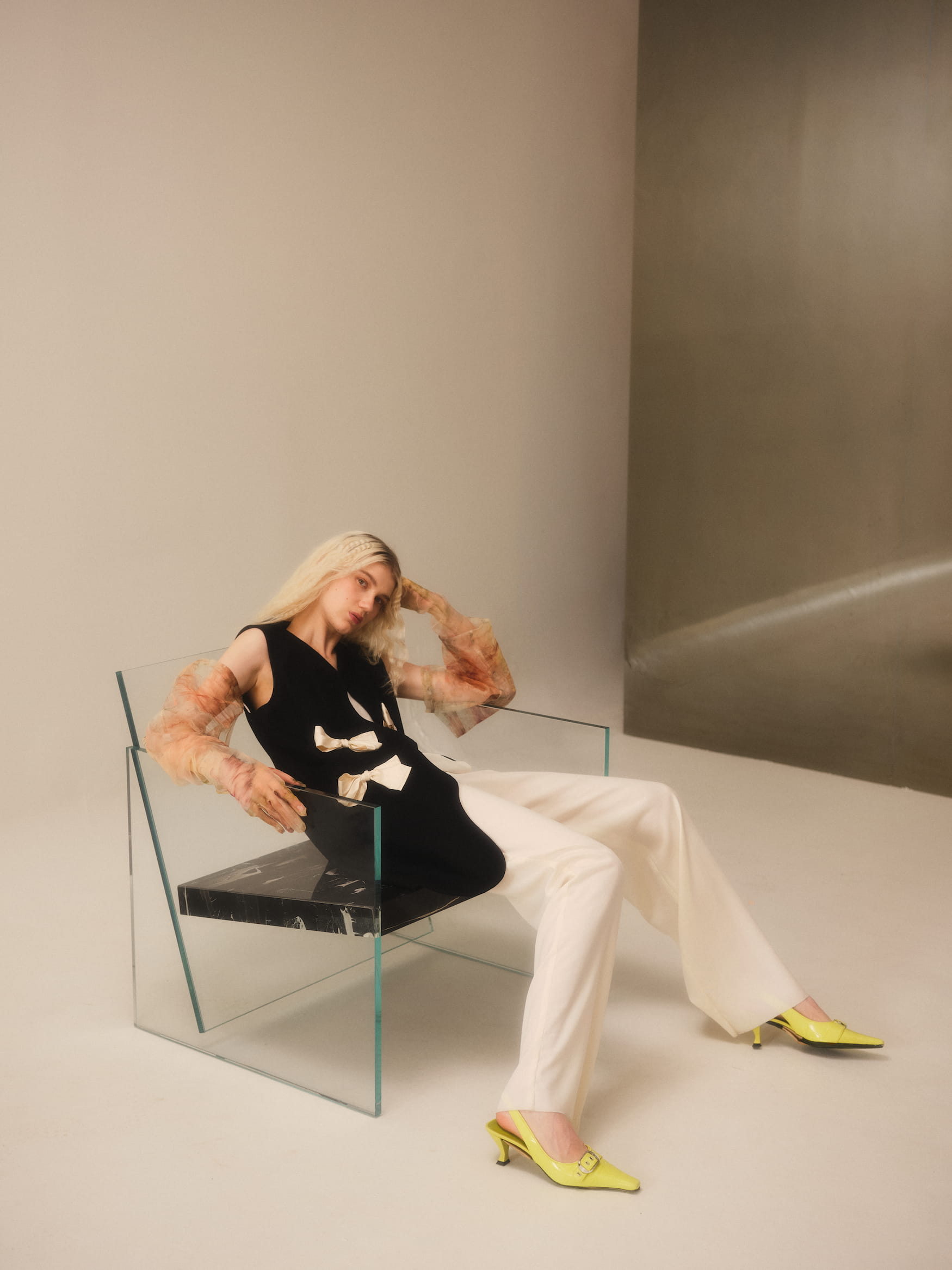 A person with long blonde hair sits casually in a modern, transparent chair, showcasing the stylish Françoise Vest by Noémiah. They're wearing a black top with semi-sheer floral sleeves, white pants tailored for a perfect fit, and lime-green heels. The minimalistic setting is accentuated by neutral tones.