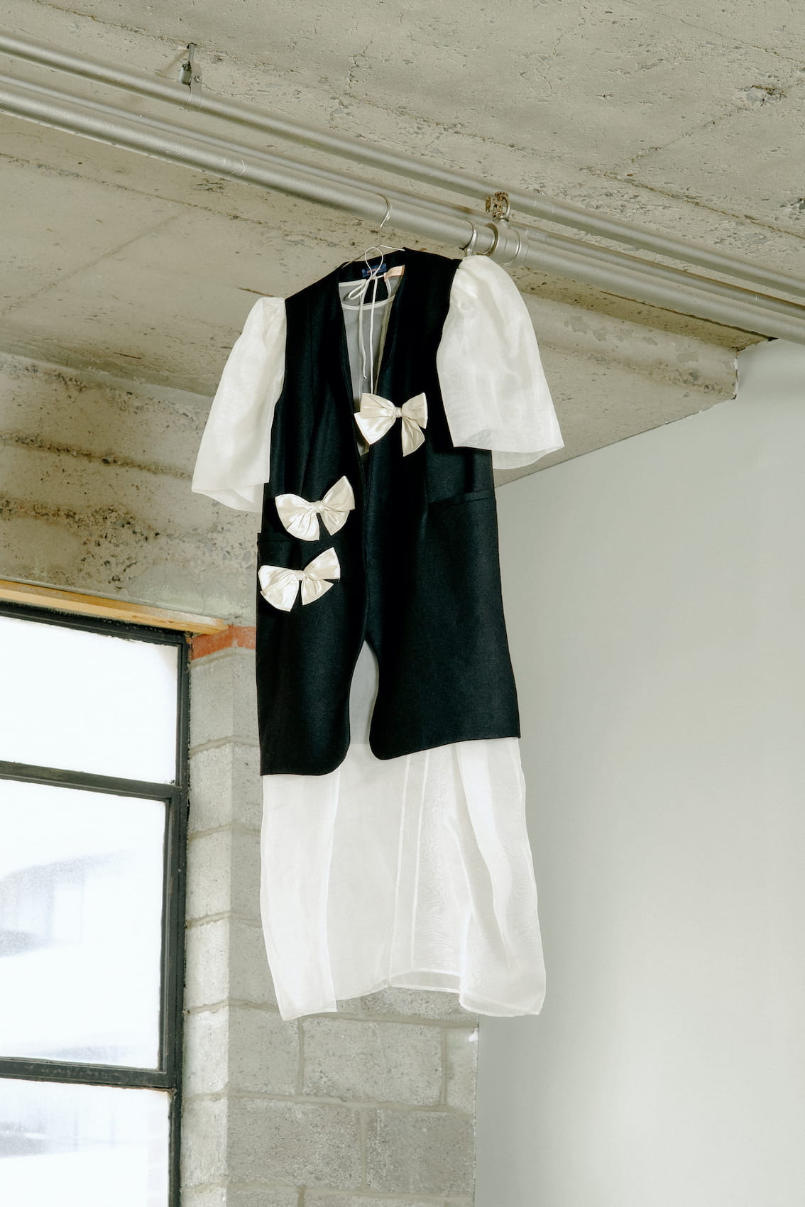 The Françoise Vest by Noémiah, a black sleeveless vest with a tailored fit and large white bows, is styled over a white dress with puffed sleeves. This ensemble is displayed hanging from a pipe near a window against a brick wall backdrop.