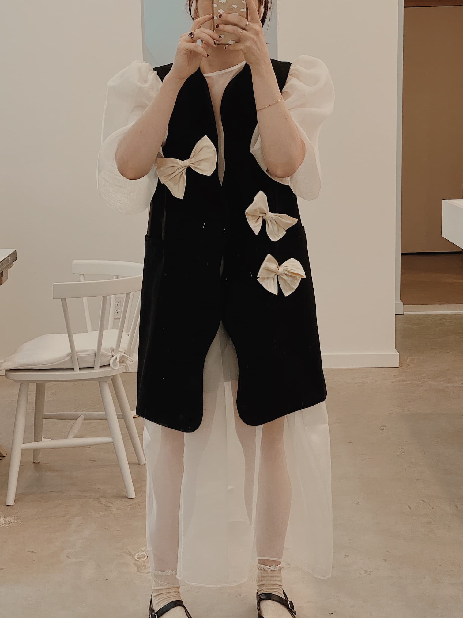 A person is taking a mirror selfie wearing the Françoise Vest by Noémiah, featuring a tailored-fit black design accented with large white bows and puffy white sleeves. They are standing in a modern room with a white chair and table in the background.