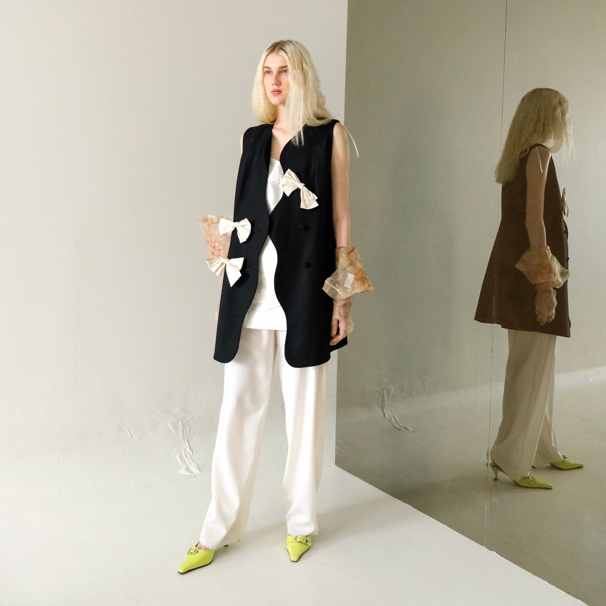 A woman with long blonde hair stands in a white room, wearing the Françoise Vest by Noémiah, which features floral bows, along with a tailored fit white outfit and bright yellow shoes. Her reflection appears in the mirror beside her.