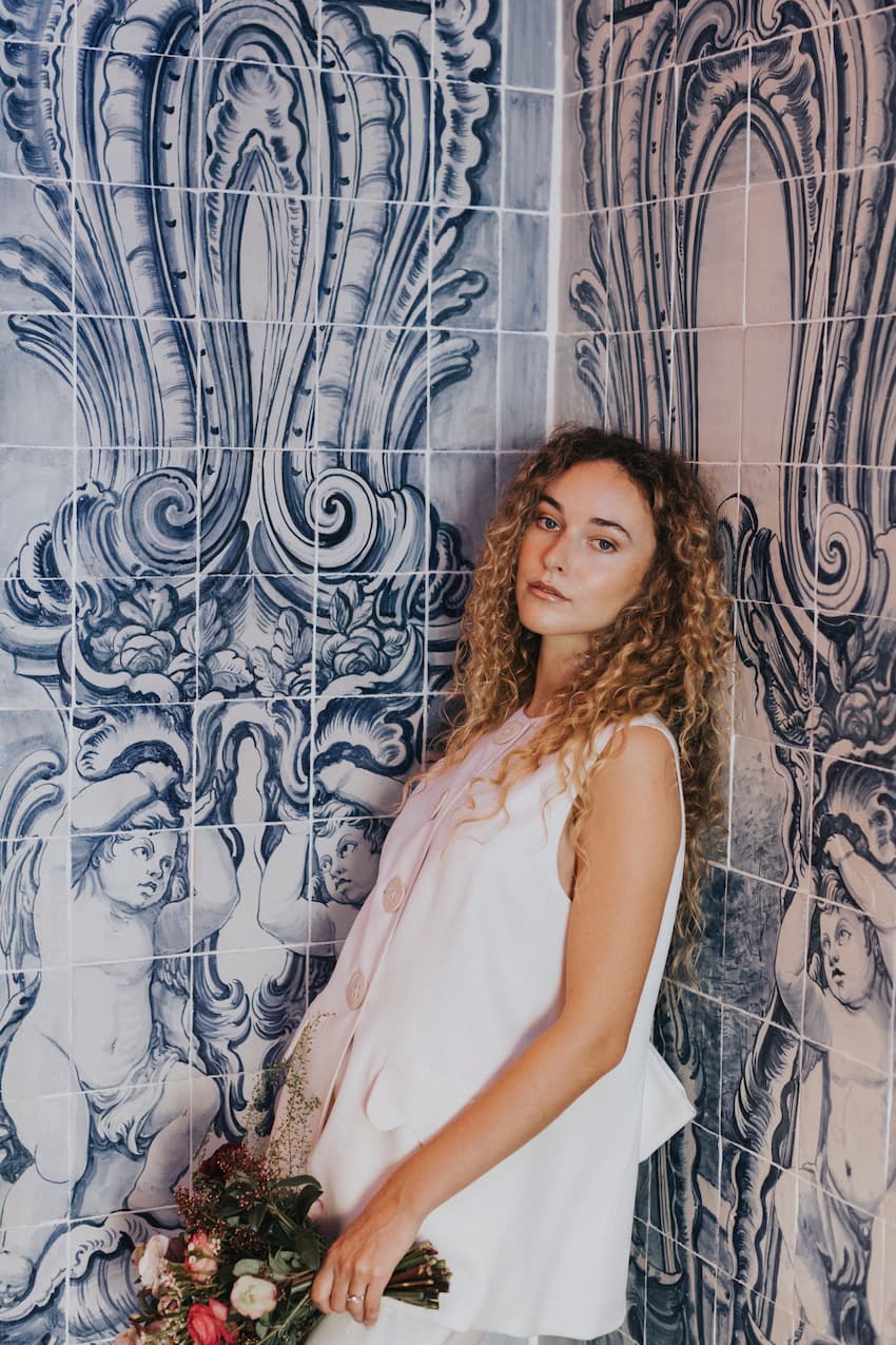 A woman with curly hair wearing the Frédérique Trousers from Noémiah's Bridal Edit and a white sleeveless top leans against a wall adorned with intricate blue and white tile art. She holds a bouquet of flowers and gazes at the camera.