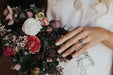 A person with long hair is holding a bouquet of mixed flowers with red, white, and pink blooms. They are wearing a white outfit that includes the stylish Frédérique Trousers — Bridal Edit from Noémiah and have a ring on their finger, suggesting a wedding or special occasion.