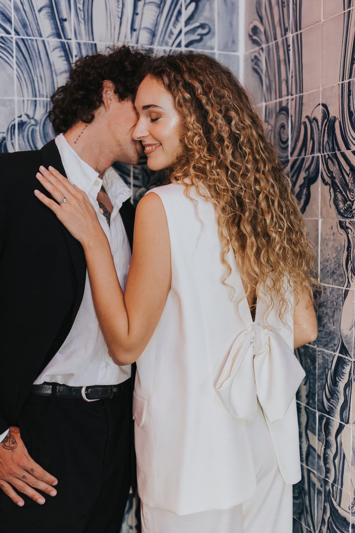 A couple stands closely in an intimate pose. The woman is wearing a white outfit with a large bow on the back from Noémiah, while the man complements his black suit with Frédérique Trousers from the Bridal Edit. They are surrounded by intricate blue and white tiled walls.