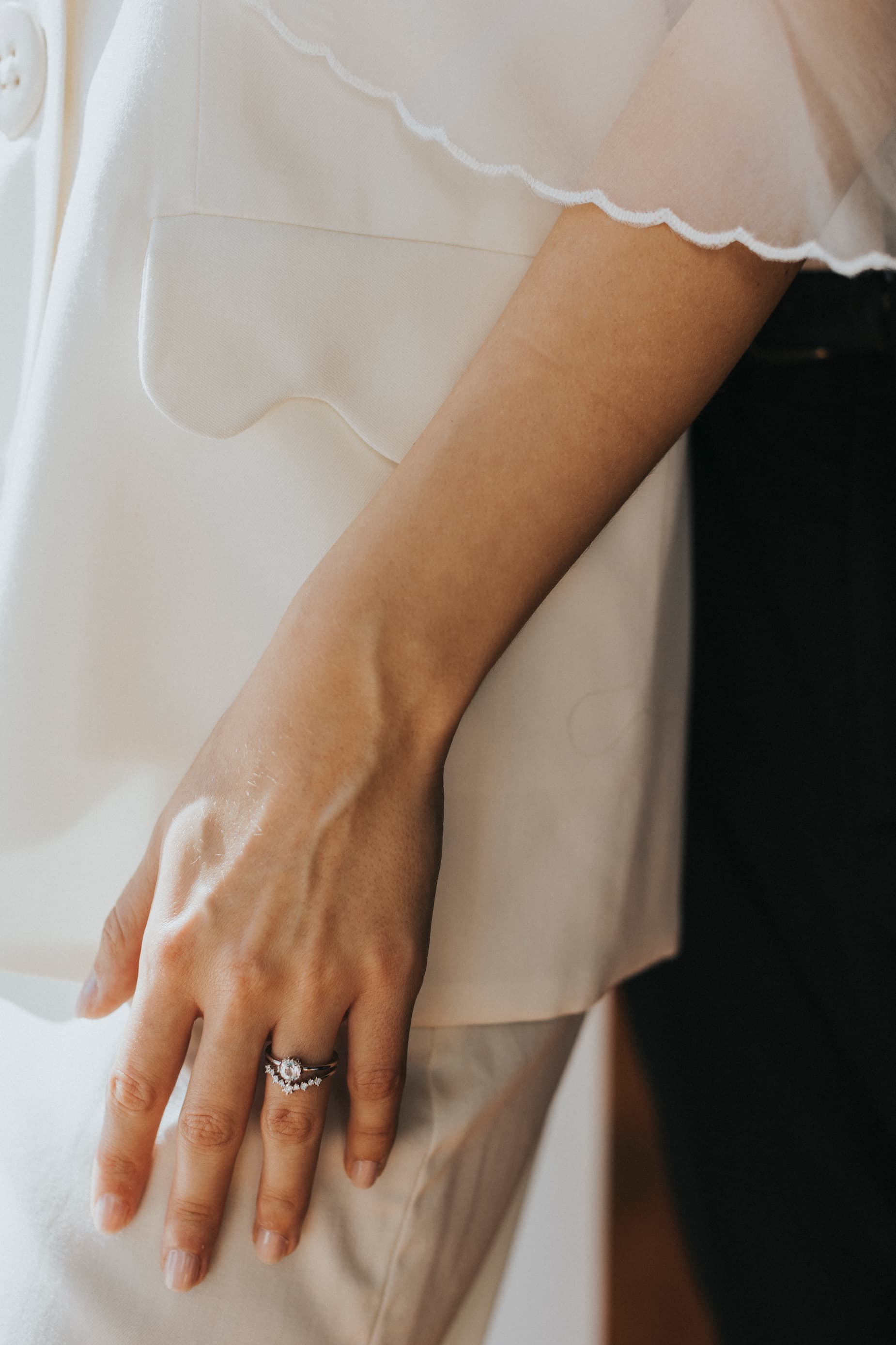 Close-up of a person in a white outfit, their left hand adorned with two rings, one featuring a prominent stone. The arm hangs relaxed, revealing the sheer fabric overlay on the sleeve, elegantly paired with Noémiah's Frédérique Trousers from the Bridal Edit that complete the chic ensemble.