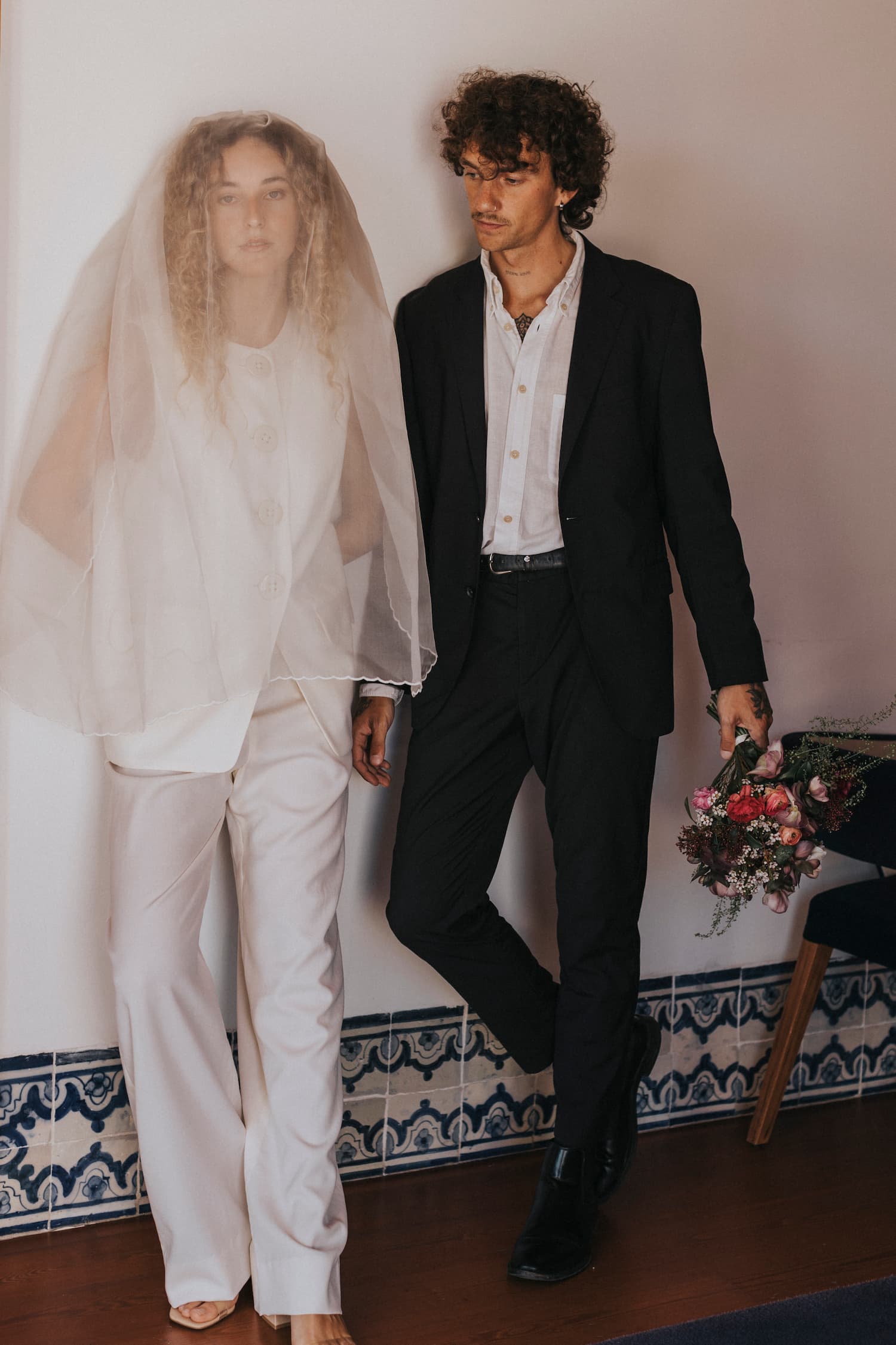 A couple stands indoors against a tiled wall. The woman, adorned in a white outfit with a veil, and the man, dressed in a dark suit complemented by Noémiah's Frédérique Trousers — Bridal Edit, hold a bouquet of flowers. Both appear relaxed and thoughtful, gazing into the distance.