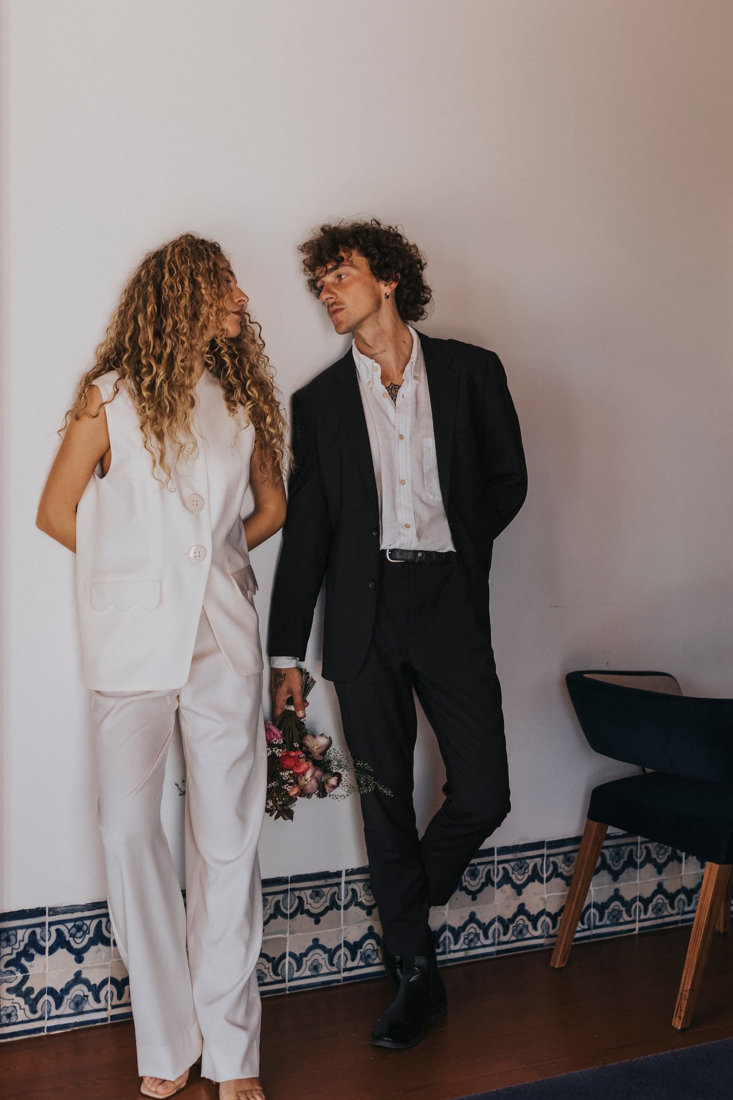 A man and woman stand indoors, facing each other. She is dressed in the Frédérique Trousers from Noémiah's Bridal Edit collection, while he wears a black suit paired with a white shirt. In his hand, he holds a bouquet of flowers. The room is adorned with decorative tiling along the wall and includes a blue chair.