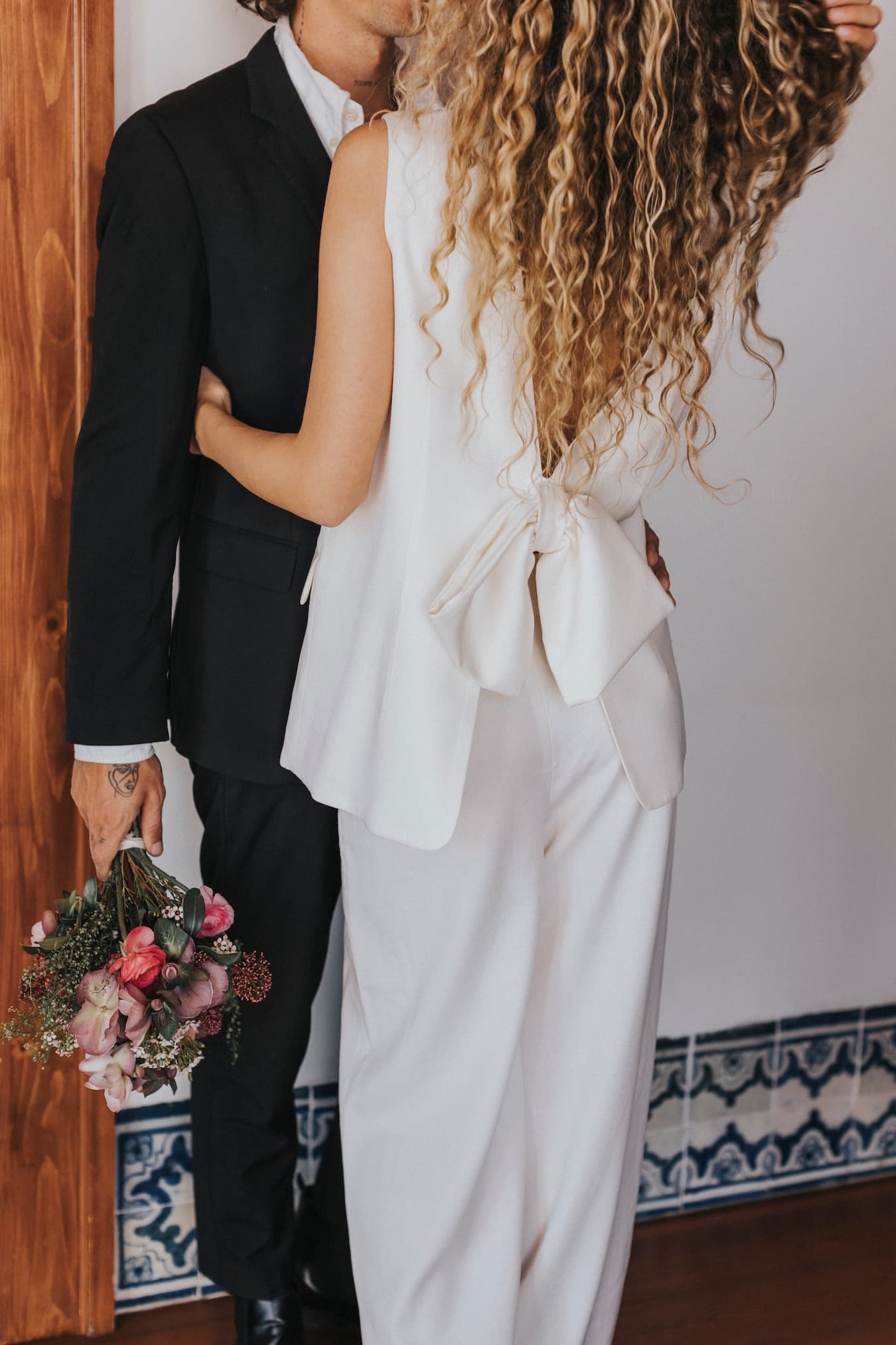 A couple stands near a patterned tile floor, embraced by a wooden wall. The woman, with long curly hair, wears an elegant white outfit adorned with a large bow. The man is dressed in a black suit and wide-leg Frédérique Trousers from the Noémiah Bridal Edit collection, holding a bouquet of pink and red flowers.