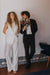 A person with curly hair in a white outfit stands next to another wearing Noémiah's Frédérique Trousers from the Bridal Edit collection, paired with a black suit and holding a bouquet of flowers. They are in a room with patterned tile decor at the base of a white wall.