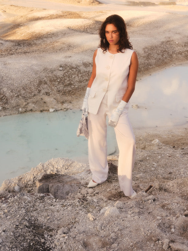 In a barren, sandy landscape near a small pool of blue water, an individual stands confidently, wearing the elegant Frédérique Trousers from Noémiah's Bridal Edit collection. Paired with gloves and holding a patterned scarf, their attire complements the soft, muted earth tones of the background.