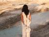 A woman in a sleeveless white dress, adorned with silver gloves, stands with her back to the camera, holding a white bag. The landscape of barren ground and a tranquil blue pool of water is bathed in warm golden light, perfectly complementing her chic Frédérique Trousers from the Bridal Edit by Noémiah.