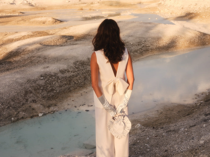 A woman in a sleeveless white dress, adorned with silver gloves, stands with her back to the camera, holding a white bag. The landscape of barren ground and a tranquil blue pool of water is bathed in warm golden light, perfectly complementing her chic Frédérique Trousers from the Bridal Edit by Noémiah.