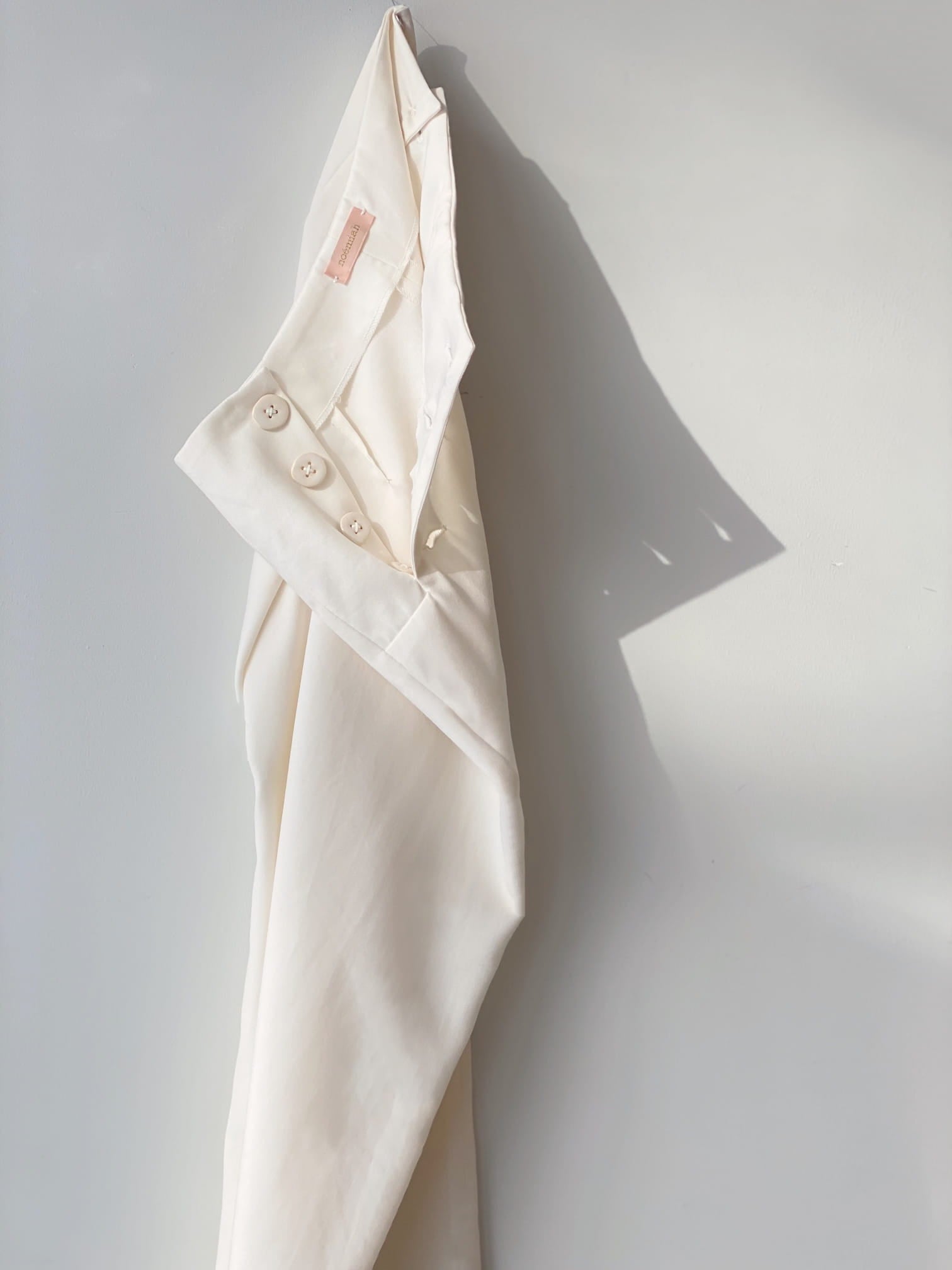 A white shirt hangs against a light gray wall, its smooth fabric accented by buttons along the sleeve. Below, the Frédérique Trousers — Bridal Edit by Noémiah rest elegantly in Merino wool twill, their texture enhanced by soft shadows, together creating a refined minimalist scene.