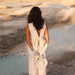 A woman in a white dress, complemented by the Frédérique Trousers from Noémiah’s Bridal Edit, stands with her back to the camera, overlooking a serene landscape with light blue water and rocky terrain. Holding a ruffled bag and wearing long gloves, she is bathed in warm, soft light.