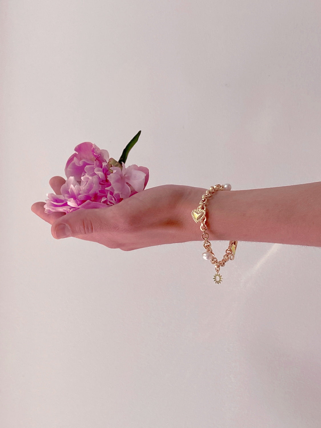 A hand adorned with the Noémiah Frida Bracelet, shimmering with charms and pearls, gently holds a pink peony against a plain background.