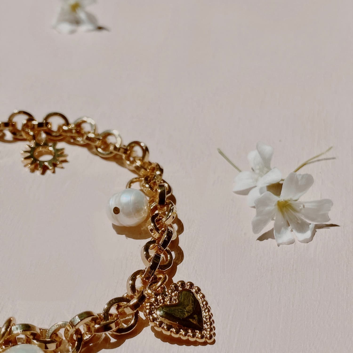 The Frida Bracelet by Noémiah, featuring a gold-plated chain adorned with a heart charm and delicate pearls, rests on a light pink surface, encircled by small white flowers.