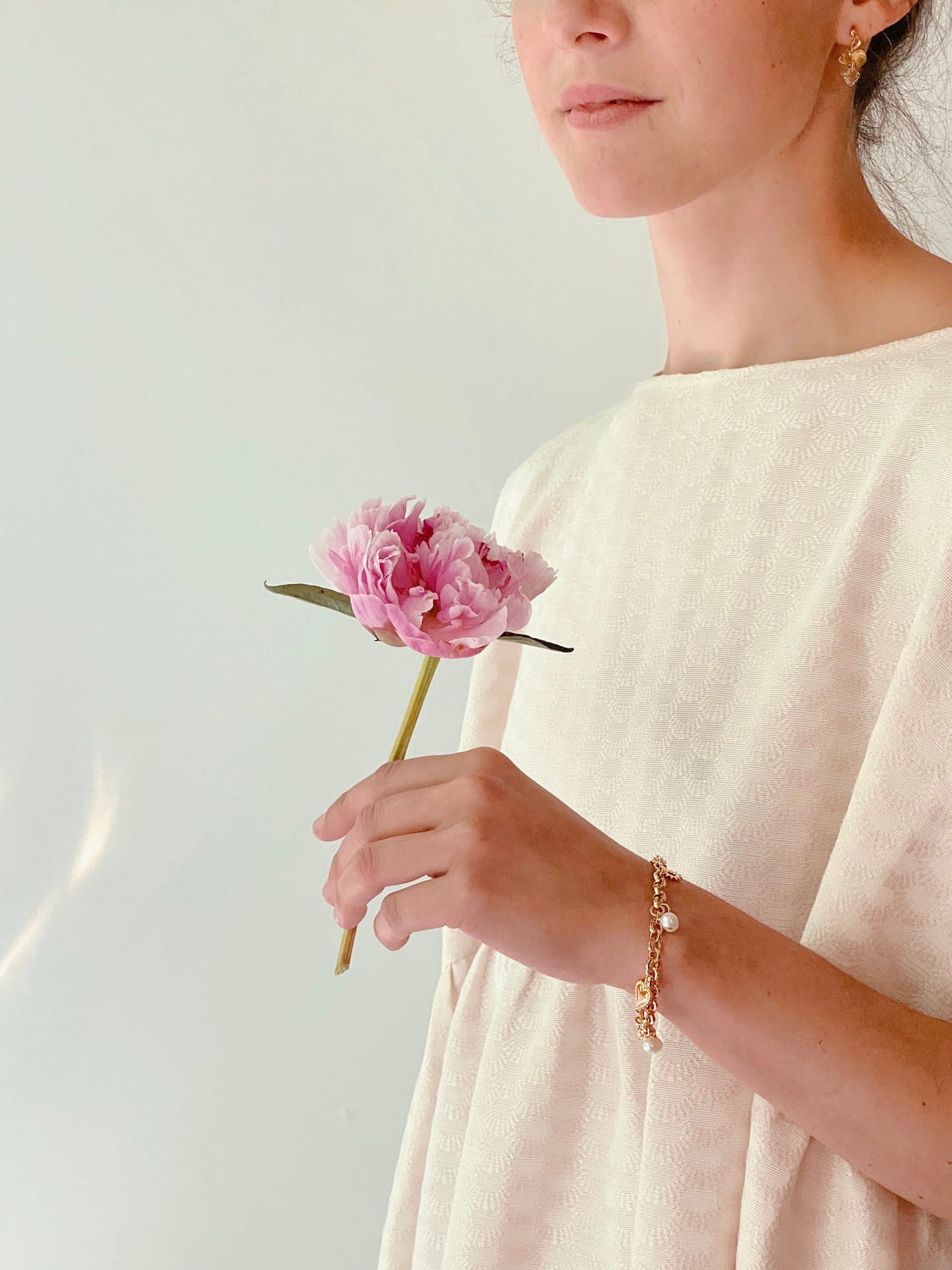 A person in a light, textured dress holds a single pink peony, adorned with the Frida Bracelet by Noémiah, which is gold-plated and set with small pearls. The background features a soft, neutral color.