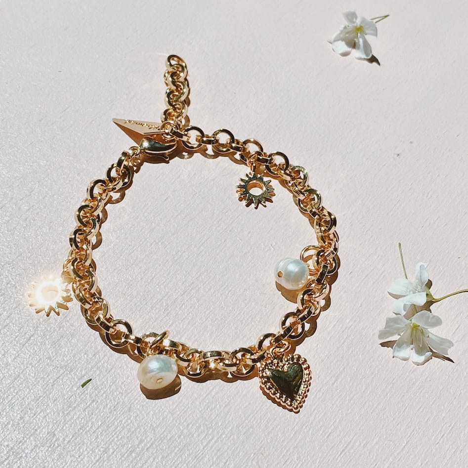 The Frida Bracelet by Noémiah, a gold-plated chain adorned with heart and flower charms and lustrous pearls, is placed on a light surface. Small white flowers are scattered around it, while the sunlight beautifully highlights the bracelet's metallic sheen and intricate charm details.