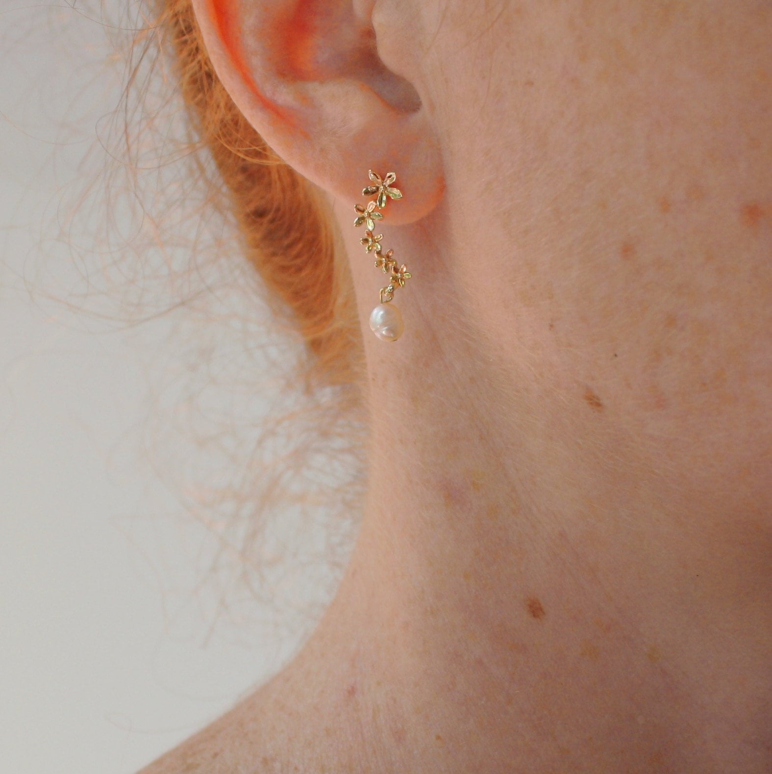 A close-up captures a person's ear graced by the Gabriella Earrings from Noémiah, featuring delicate, tarnish-resistant flower designs and adorned with a small freshwater pearl. The elegant, vine-like design enhances the beauty of the ear, framed by light-colored hair and charming freckles.