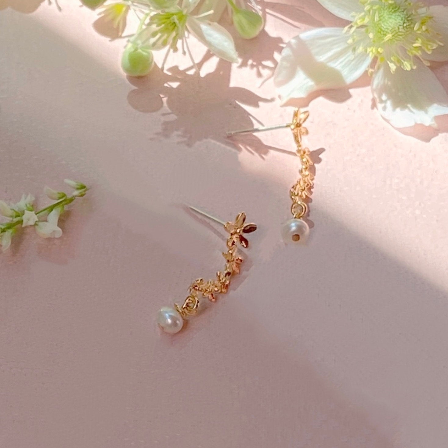 The Gabriella Earrings by Noémiah, featuring delicate gold flowers with tarnish-resistant properties and elegant pearl drops, are showcased on a soft pink surface surrounded by small white blossoms.