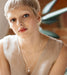 A person with short blond hair and light eyebrows sits in a softly lit interior, wearing a sleeveless beige top, the Gabriella Earrings by Noémiah, and a tarnish-resistant silver necklace with a pendant. A large green plant is visible in the blurred background.