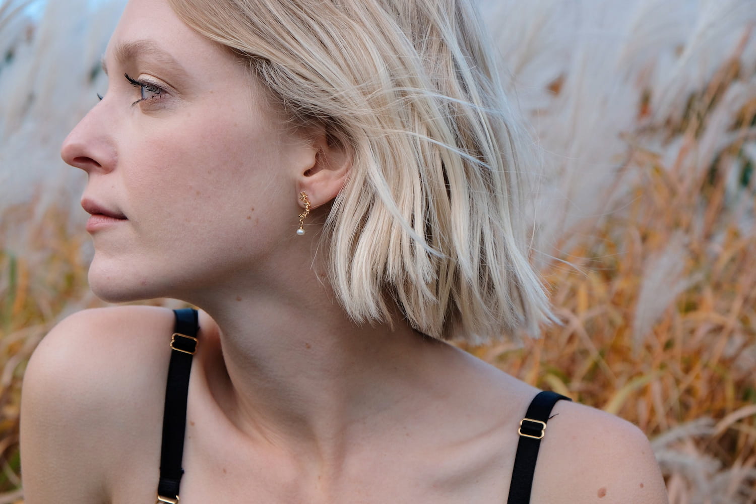 A person with short blond hair gazes sideways in an outdoor setting surrounded by tall grasses. They wear a black strap top and the tarnish-resistant Gabriella Earrings from Noémiah. The background features blurred beige and green foliage.