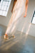 A person in the Gia Dress by Noémiah, made from long, translucent champagne silk organza, gracefully walks across a sunlit room with expansive windows. White heeled sandals are visible on their feet as the floor captures reflections of light. The slightly blurred focus lends a soft effect, ideal for non-traditional brides.