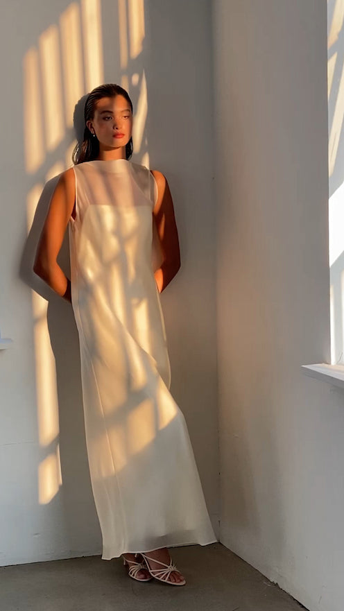 Wearing a Noémiah Gia Dress, an individual stands against a wall bathed in dramatic shadowed sunlight. Their hair is elegantly pulled back, and they complement the look with strappy sandals. The scene embodies a calm and reflective mood, perfectly capturing the essence of non-traditional brides.