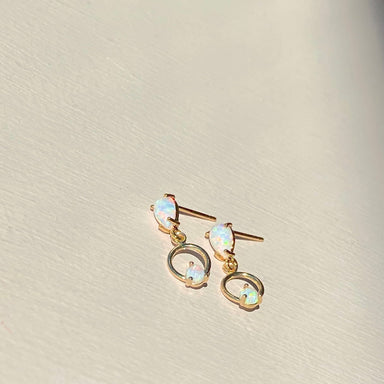 Introducing the Gigi Earrings by Noémiah: a stunning pair of gold opal teardrop earrings. These exquisite earrings feature small opal studs and larger dangling opals, elegantly encircled by a gold ring. Set against a light background, the opals radiate with a subtle play of colors that evoke the delicate beauty of butterfly wings fluttering in sunlight.