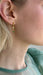 Close-up of a person with light skin and blonde hair wearing Noémiah’s Hélène Earrings—minimalist drop earrings featuring a small emerald green gemstone. The person is dressed in a sheer, pale green top.