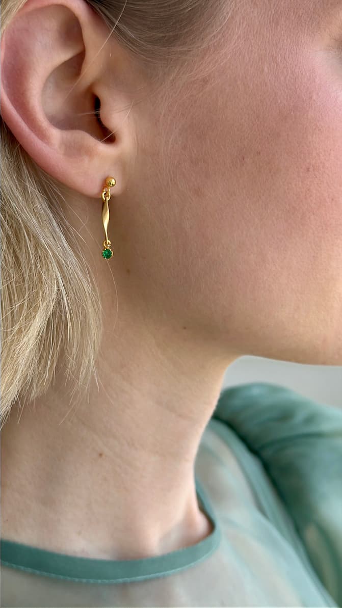 Close-up of a person with light skin and blonde hair wearing Noémiah’s Hélène Earrings—minimalist drop earrings featuring a small emerald green gemstone. The person is dressed in a sheer, pale green top.