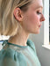A woman with short blonde hair, shown in profile by a sunlit window, wears minimalist Hélène Earrings from Noémiah and a sheer light green top.