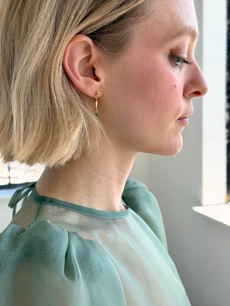 A woman with short blonde hair, shown in profile by a sunlit window, wears minimalist Hélène Earrings from Noémiah and a sheer light green top.
