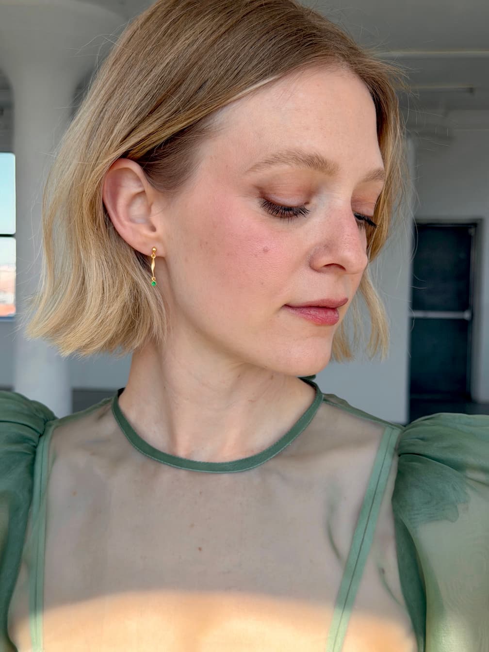 A person with short blonde hair wearing a sheer green top and Noémiah’s Hélène Earrings looks down and to the side in a bright room with white walls and large windows.