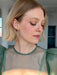 A person with short blonde hair wearing a sheer green top and Noémiah’s Hélène Earrings looks down and to the side in a bright room with white walls and large windows.