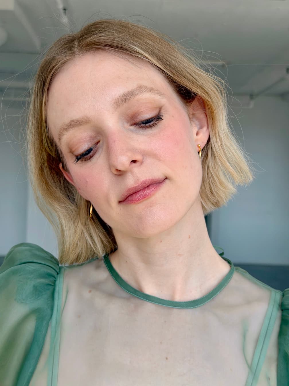A woman with short blonde hair and light makeup wears the Hélène Earrings by Noémiah with a sheer green top, looking down serenely in a softly lit indoor setting.