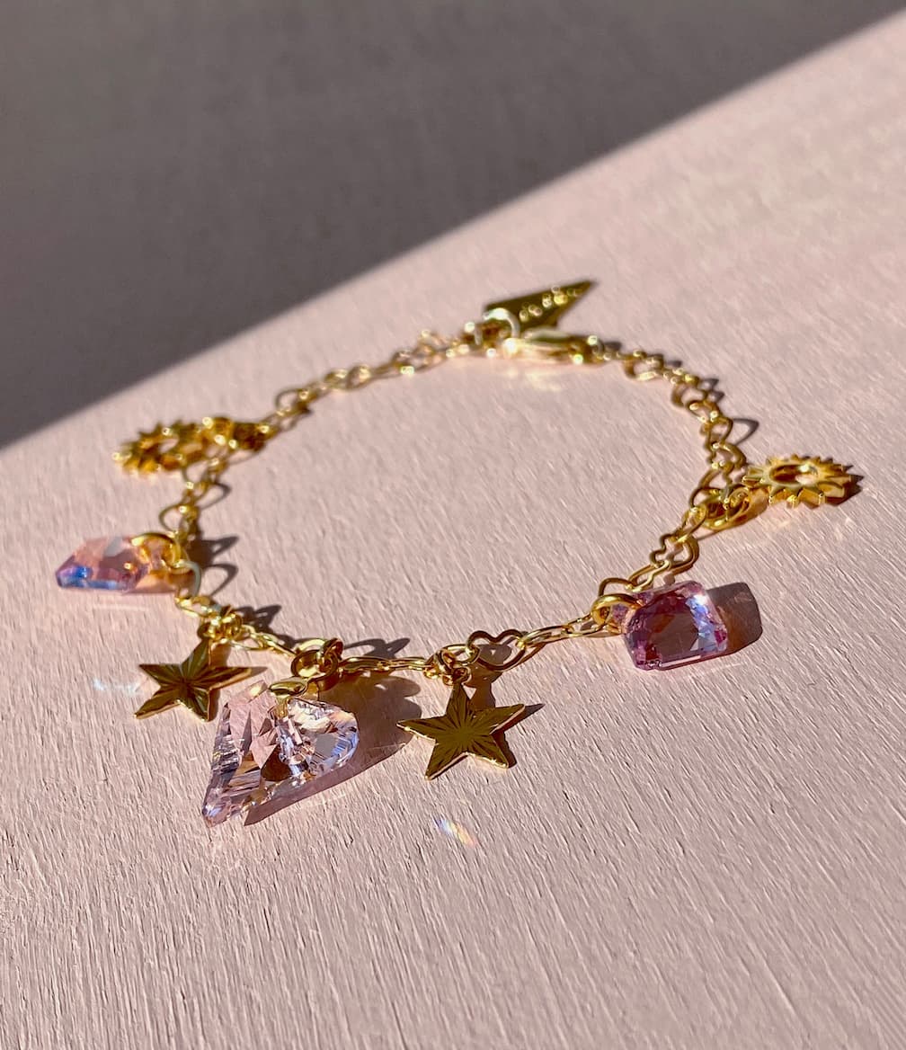 The Henriette Bracelet by Noémiah, with its gold-plated charm and adorned with Swarovski crystals, star-shaped, and gemstone pendants, rests on a light pink surface. Illuminated by sunlight, it casts soft shadows that highlight the elegance of its design.