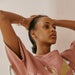 A woman in a pink blouse adorned with gold embroidery sits with her hands behind her head, exuding calm. She accentuates her look with gold hoop earrings and the Henriette Bracelet by Noémiah, while a heart chain sparkles subtly against the soft, light pink wall.