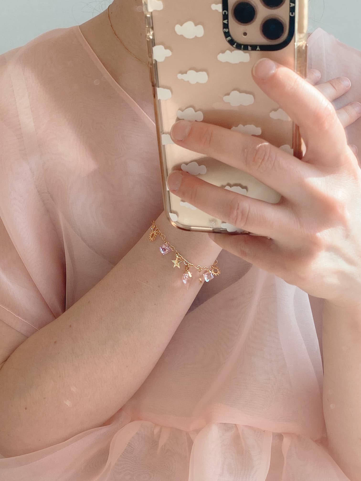 A person in a sheer pink blouse holds a phone with a cloud-patterned case. Around their wrist is the Henriette Bracelet by Noémiah, adorned with pink Swarovski crystals and clear gems, adding a touch of sparkle.